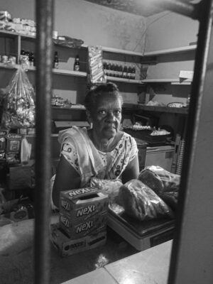 A female shopkeeper in a small grocery store surrounded by products, captured in black and white.